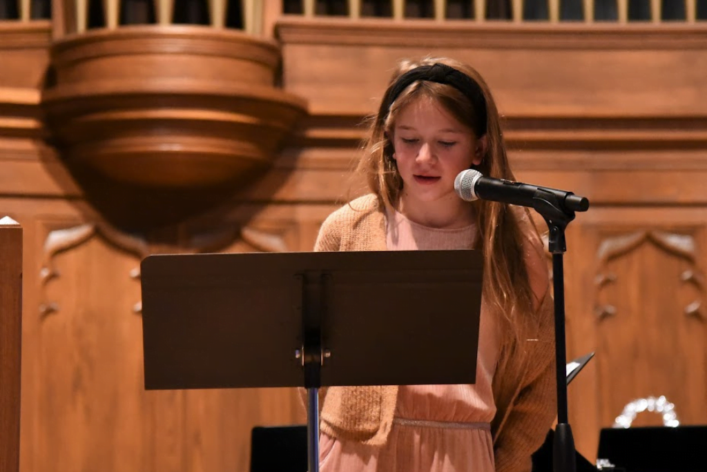 Young girl wearing peach colored dress and black headband speaking into a microphone at the front of a church.